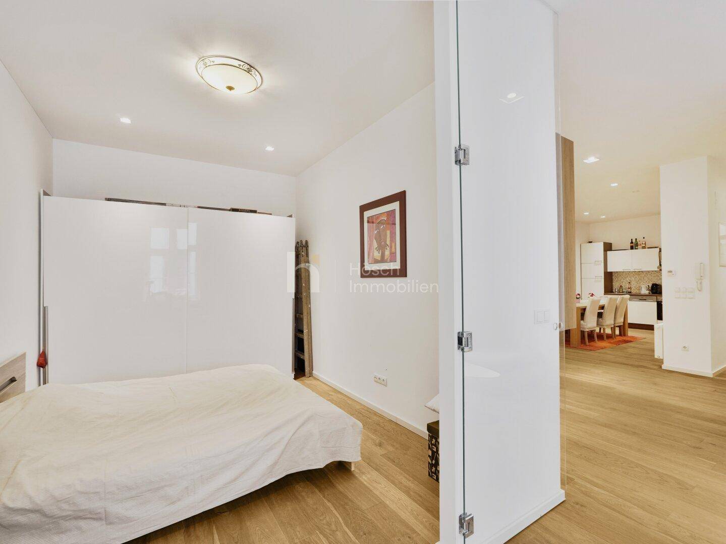 Schlafzimmer mit Doppelbett, weißem Kleiderschrank und Parkettboden, offen zum Wohnbereich.