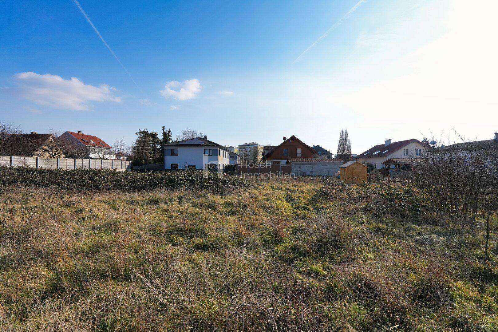 Offenes Feld mit Blick auf mehrere Wohnhäuser und einen kleinen Schuppen unter klarem Himmel.