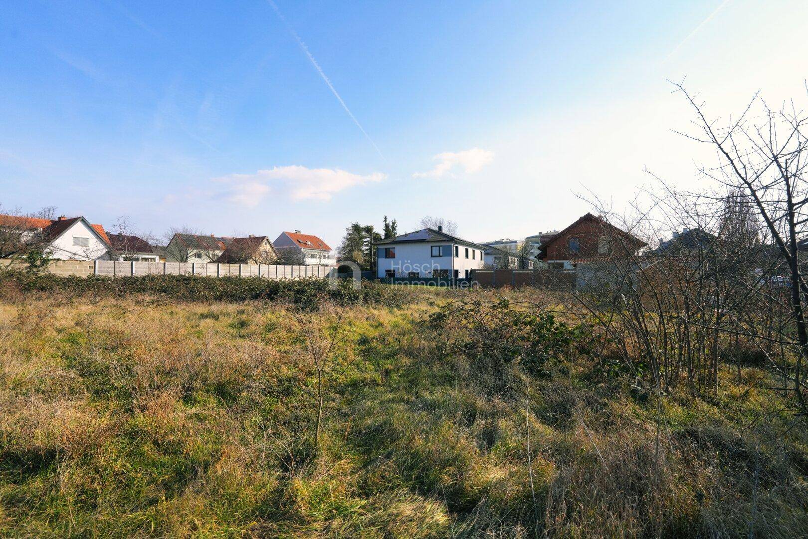 Weitläufiges Grundstück mit trockener Grasfläche und Häusern in der Ferne unter blauem Himmel.