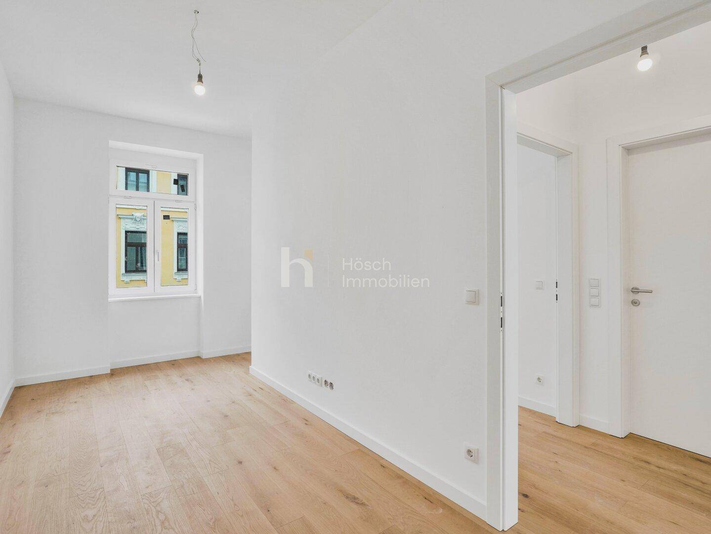Helles, leeres Zimmer mit Holzboden und einem Fenster, das Tageslicht hereinlässt.
