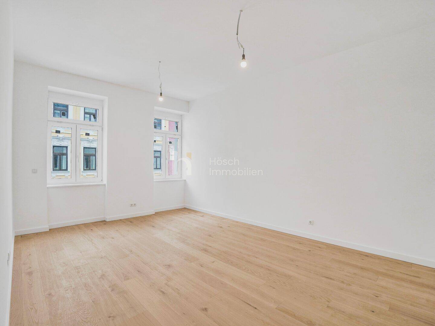Geräumiges, helles Zimmer mit Holzboden und zwei großen Fenstern, ideal für Wohnzwecke.