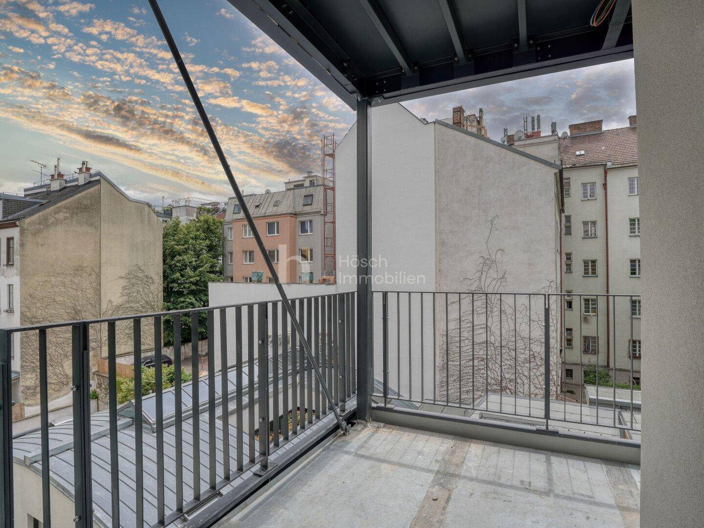 Balkon mit Metallgeländer und weitem Blick über die Dächer der Stadt bei Sonnenuntergang.