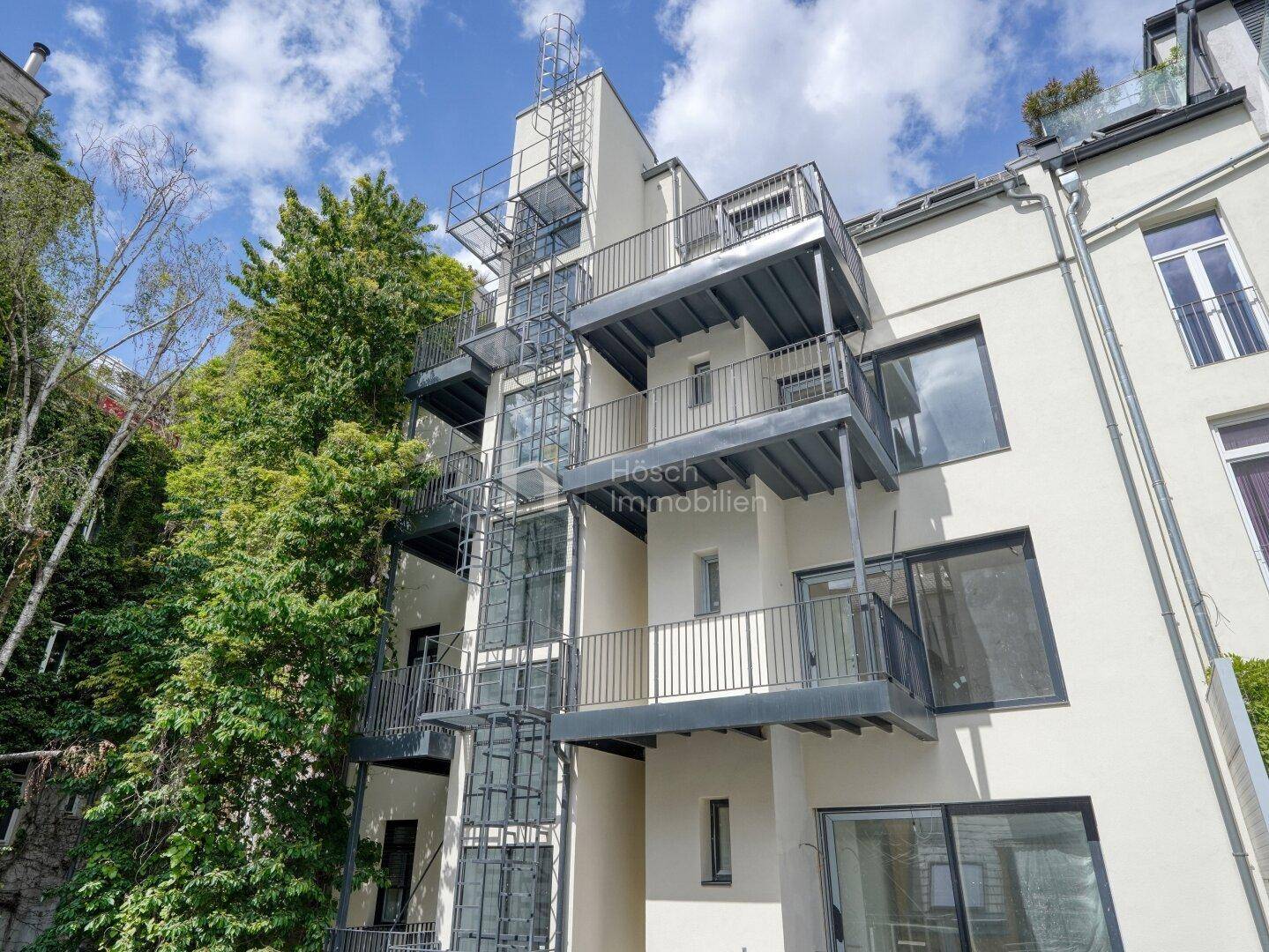 Moderne Gebäudefassade mit mehreren Balkonen und einem externen Aufzug, umgeben von Bäumen.
