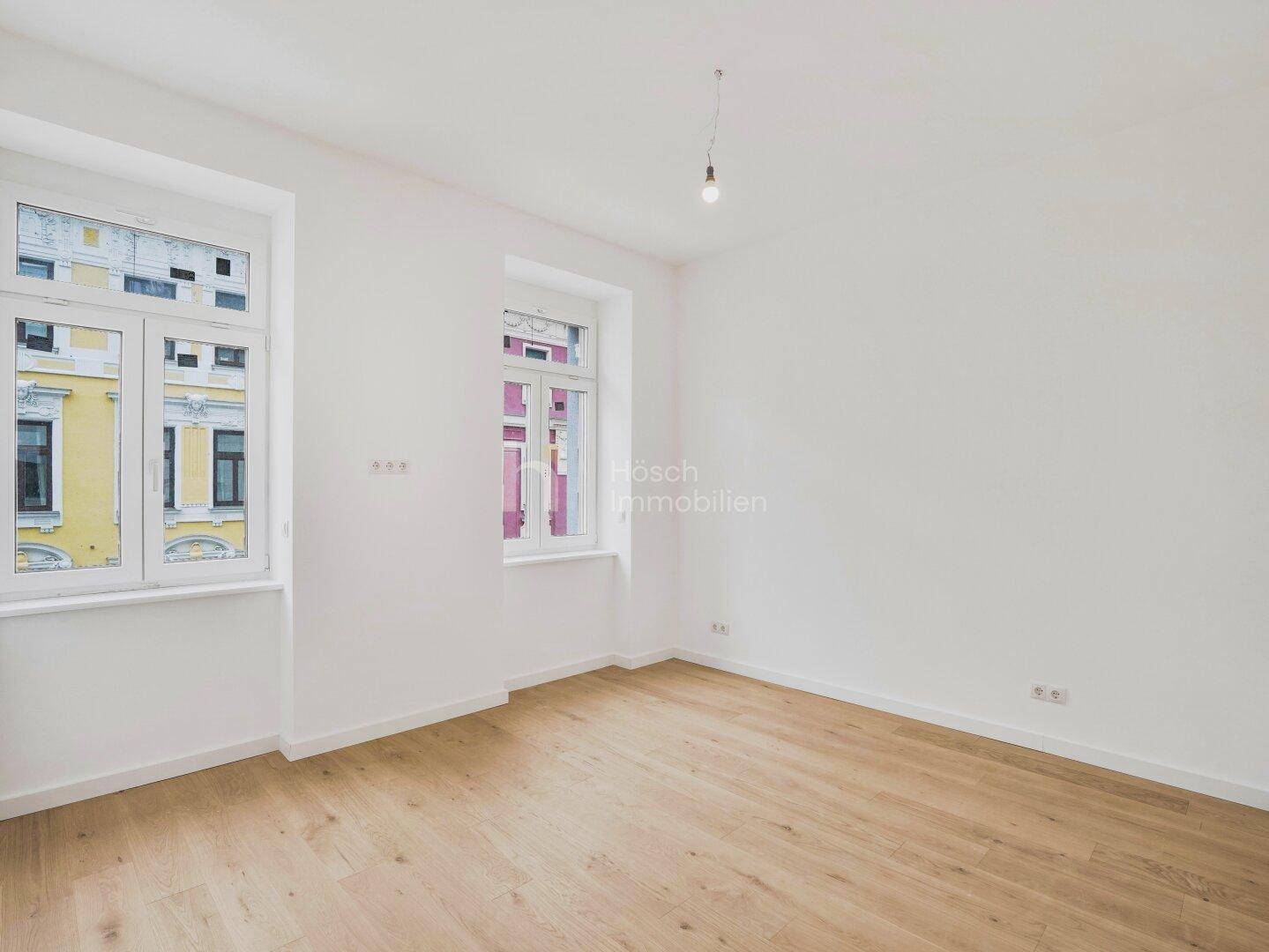 Lichtdurchflutetes Zimmer mit Holzfußboden und zwei großen Fenstern zur Straße.