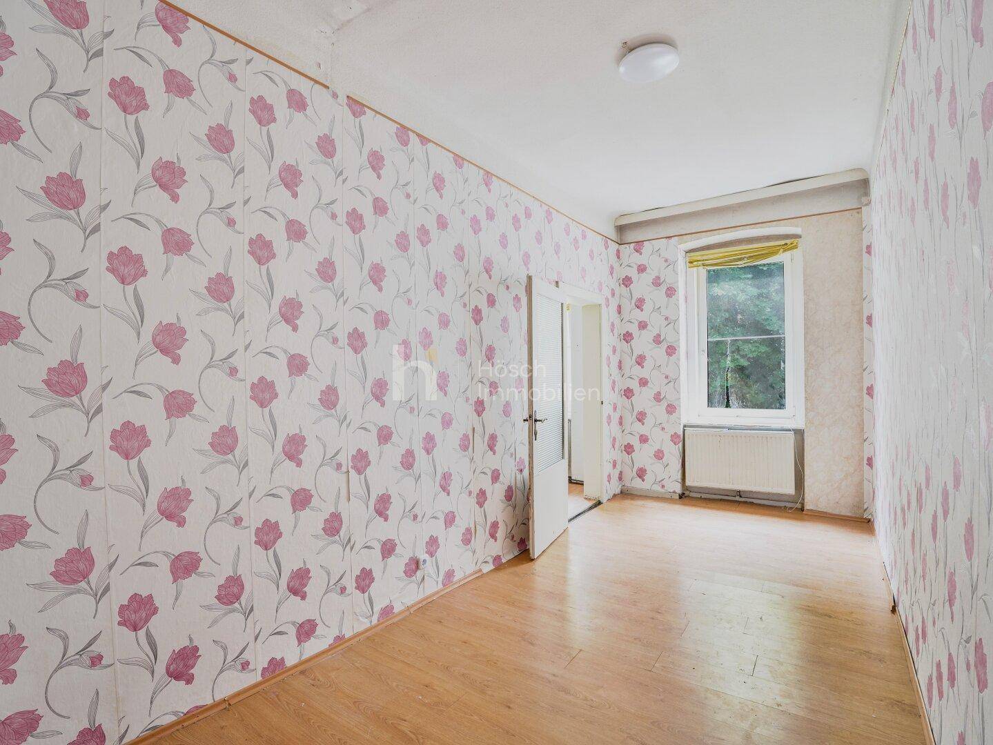 Gemütliches Zimmer mit floraler Tapete, Holzboden und einem Fenster mit Blick ins Grüne.