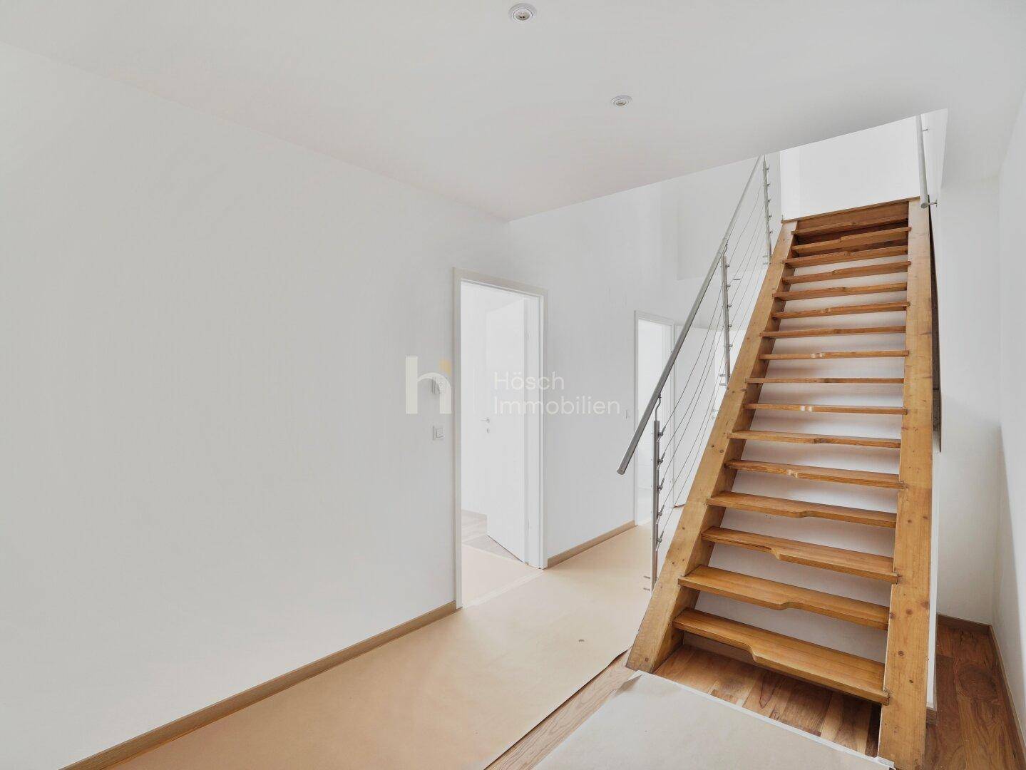 Heller Flur mit moderner Holztreppe und Edelstahlgeländer, der zu weiteren Räumen führt.