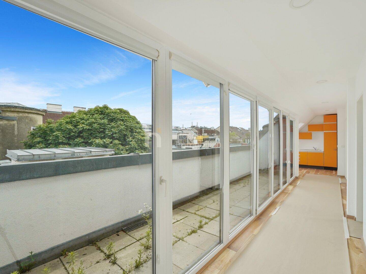Heller Innenraum mit großen Fensterfronten und Blick auf die Stadt, im Hintergrund eine kleine Küchenzeile.