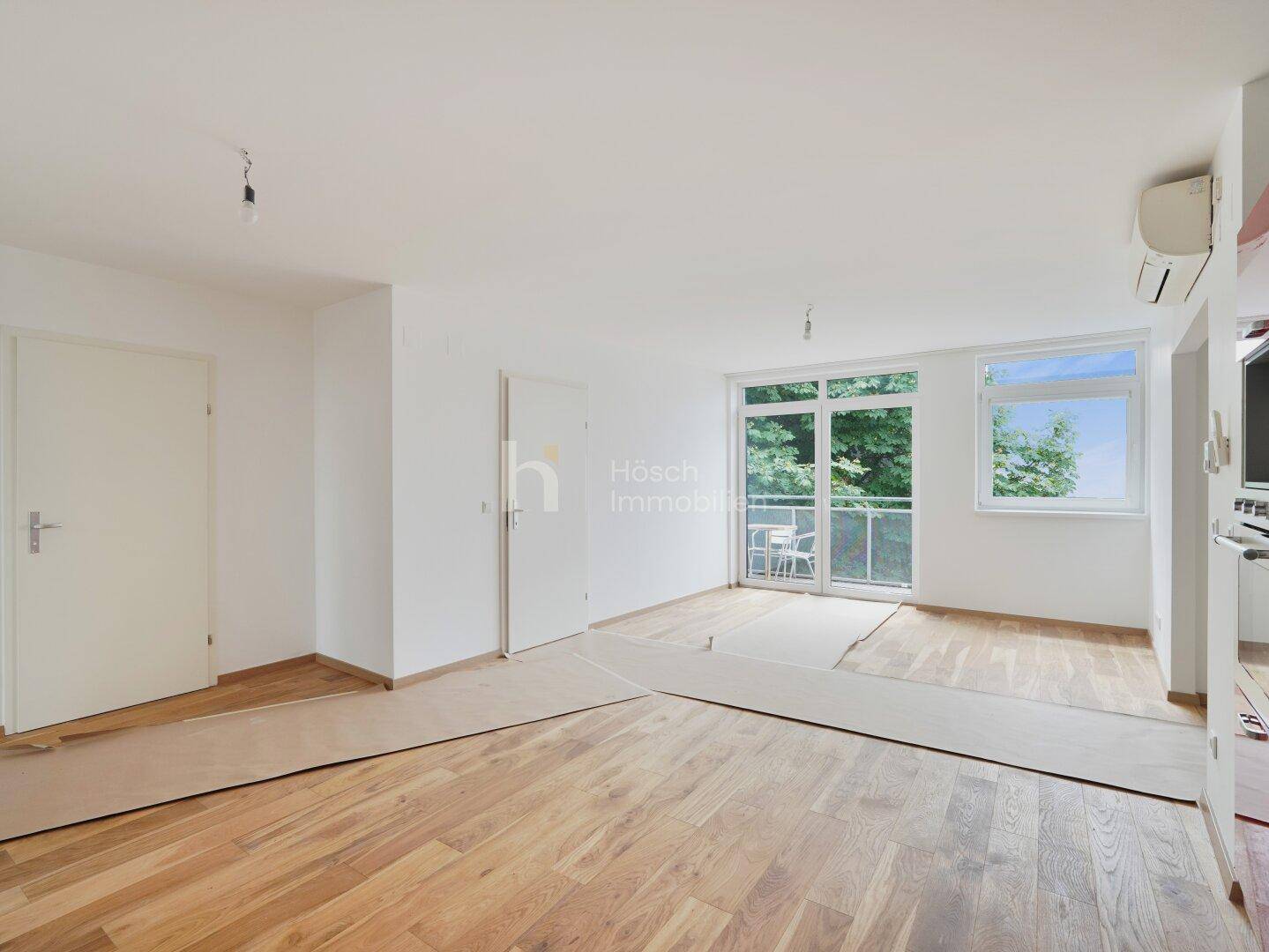 Heller Wohnraum mit Holzboden, Klimaanlage und Zugang zu einem Balkon mit Blick ins Grüne.