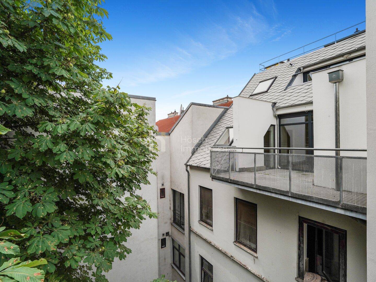 Blick auf einen Balkon mit Metallgeländer und umliegende Gebäude mit viel Grün.