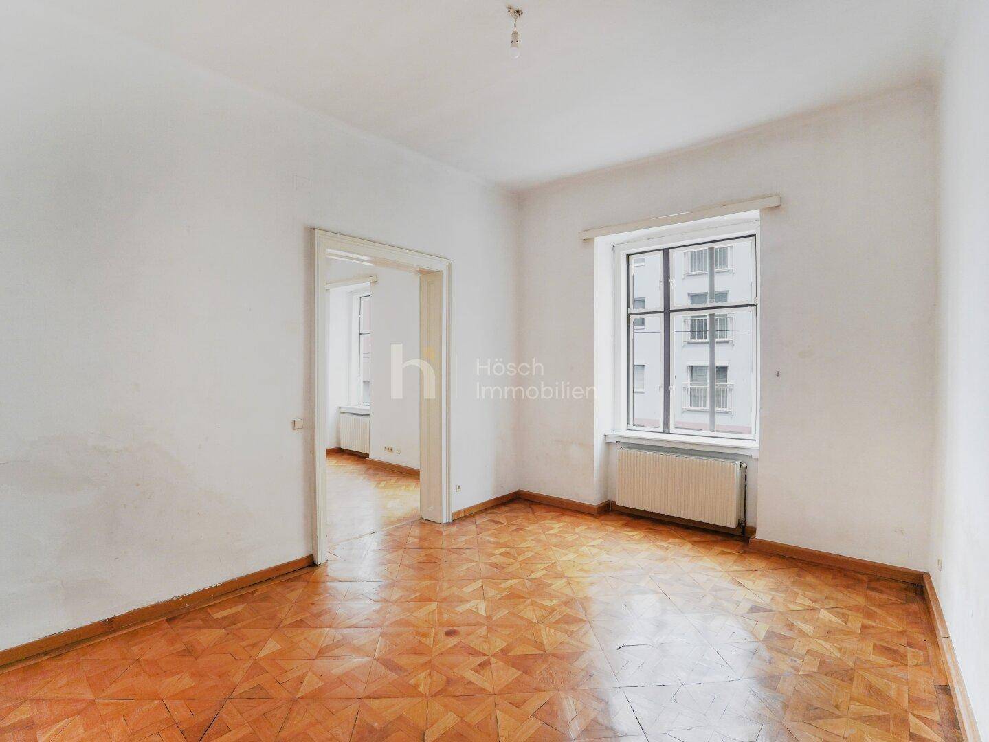 Großes Zimmer mit traditionellem Fischgrätparkett und einem Fenster, das den Raum mit Tageslicht versorgt.