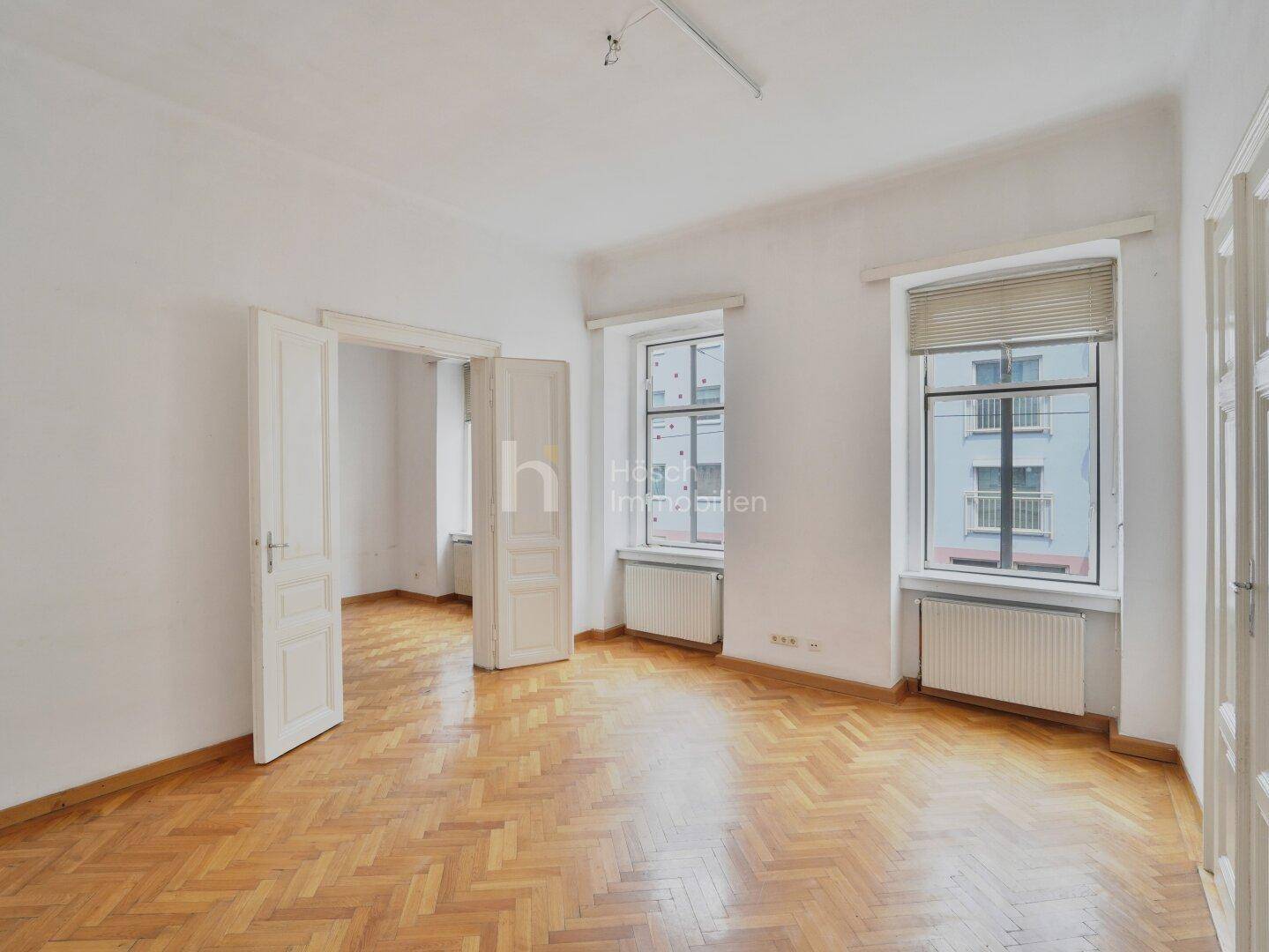 Geräumiges Zimmer mit Fischgrätparkett, hohen Decken und zwei Fenstern, die viel Licht hereinlassen.