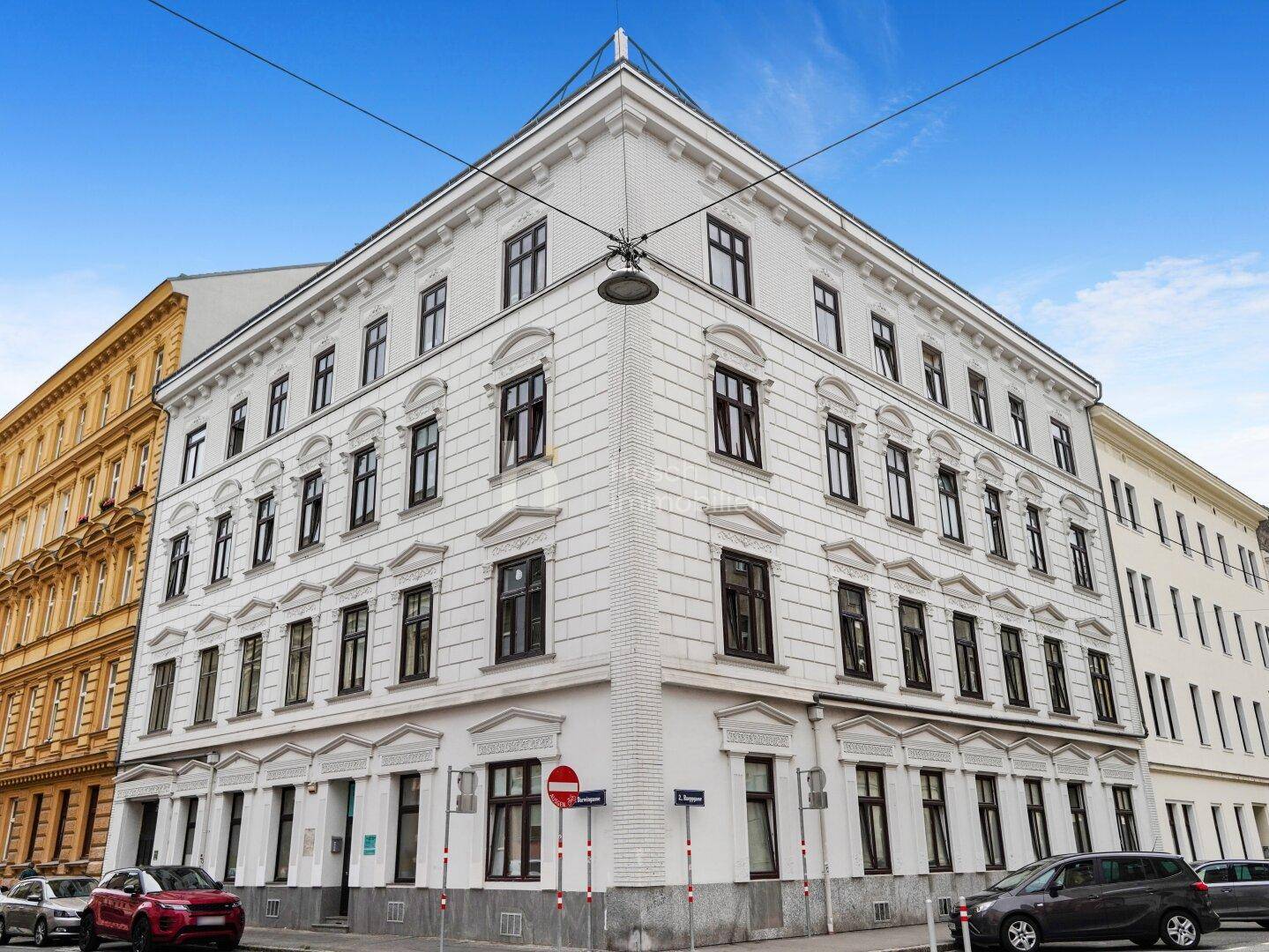 Elegantes weißes Eckgebäude mit klassischer Architektur und vielen Fenstern in einer städtischen Umgebung.