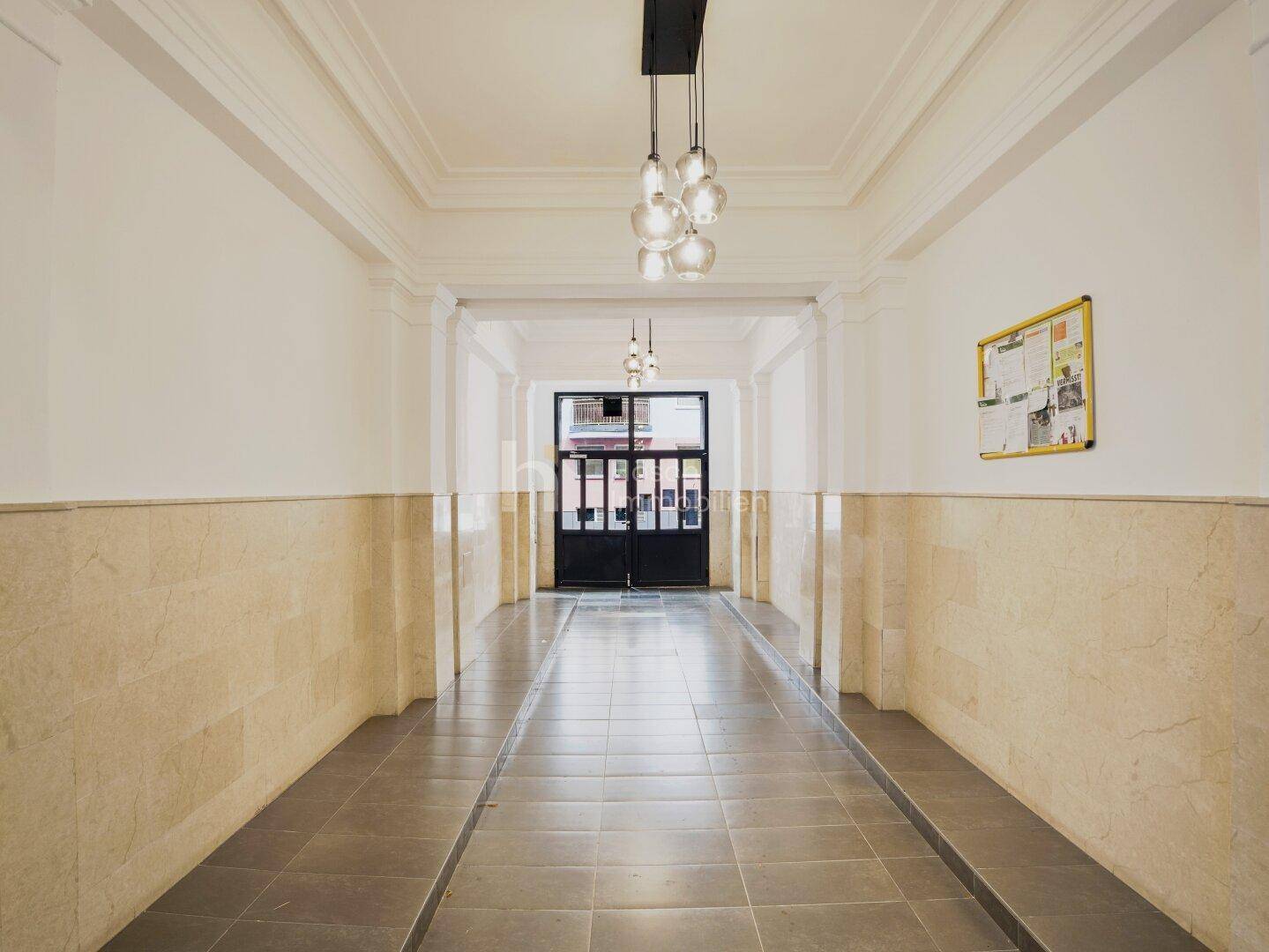 Langer, heller Eingangsbereich eines Gebäudes mit Fliesenboden und modernen Deckenleuchten.