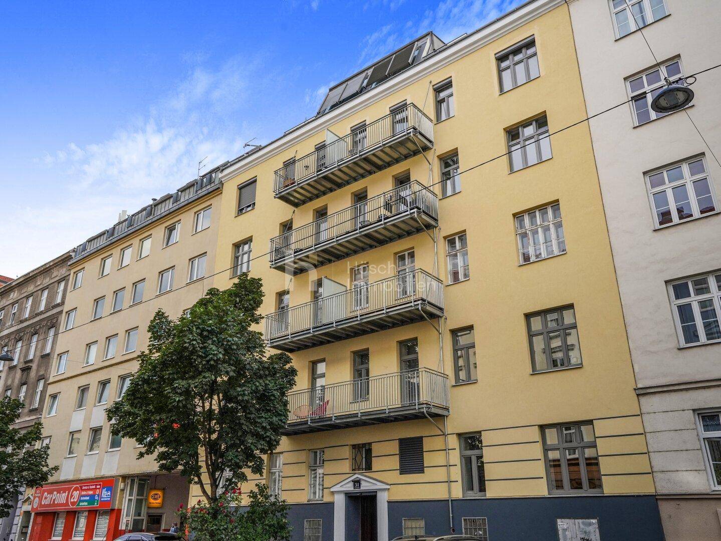 Ansicht der gelben Fassade eines Mehrfamilienhauses mit mehreren Balkonen und Geschäften im Erdgeschoss.