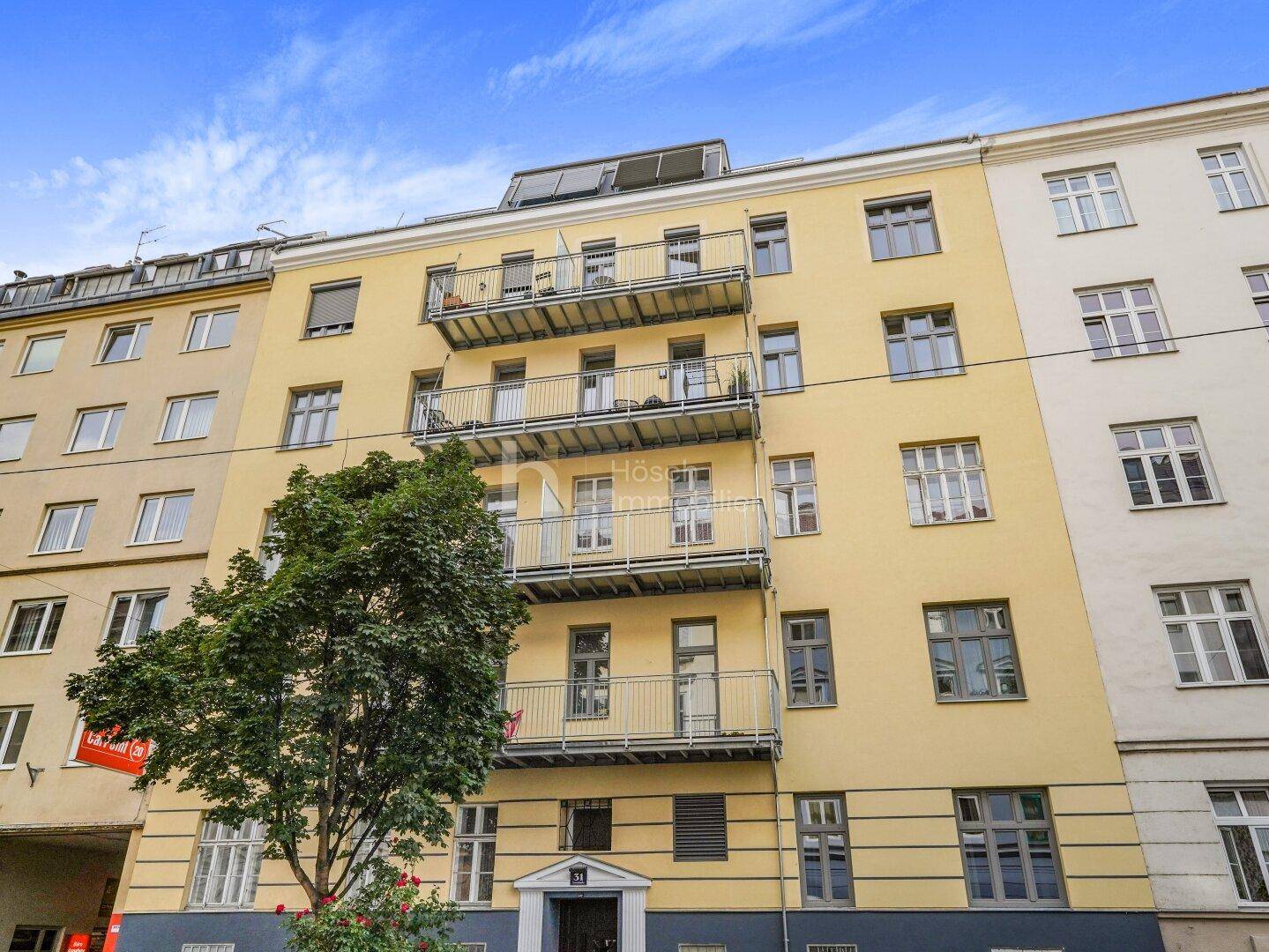 Ansicht der gelben Fassade eines Mehrfamilienhauses mit mehreren Balkonen unter blauem Himmel.