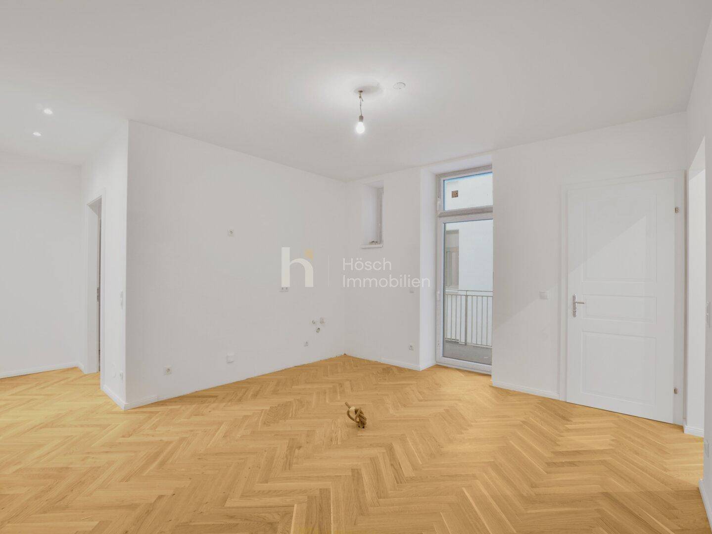 Heller Raum mit Fischgrätparkett, weiß gestrichenen Wänden und Zugang zu einem Balkon.