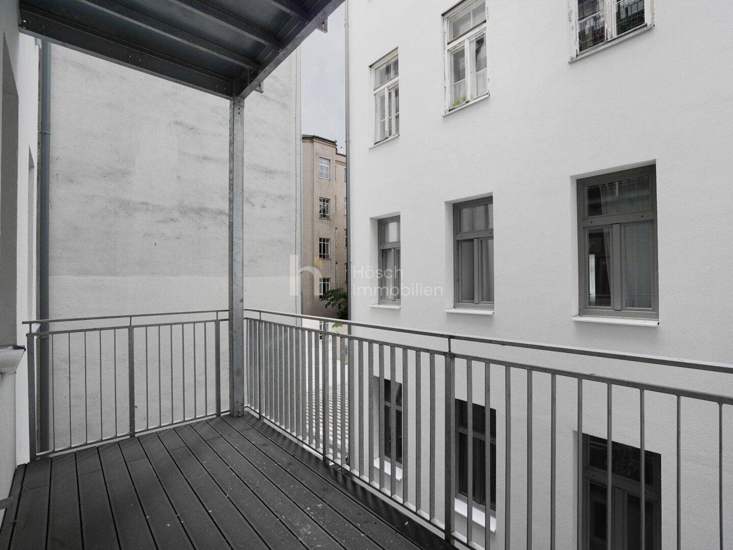Kleiner Balkon mit Holzboden und Metallgeländer, umgeben von weißen Gebäudefassaden.