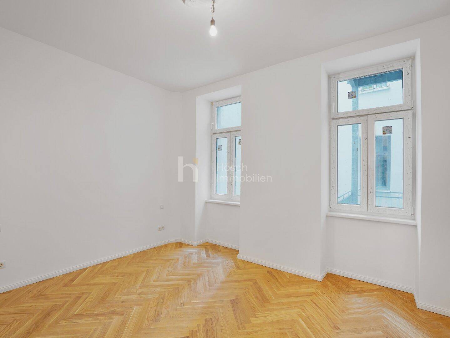 Geräumiges Zimmer mit Fischgrätparkett und zwei großen Fenstern, die viel Licht hereinlassen.