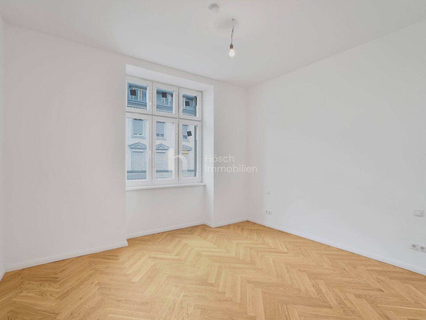 Helles Zimmer mit Fischgrätparkett und einem großen Fenster, das auf die Straße blickt.
