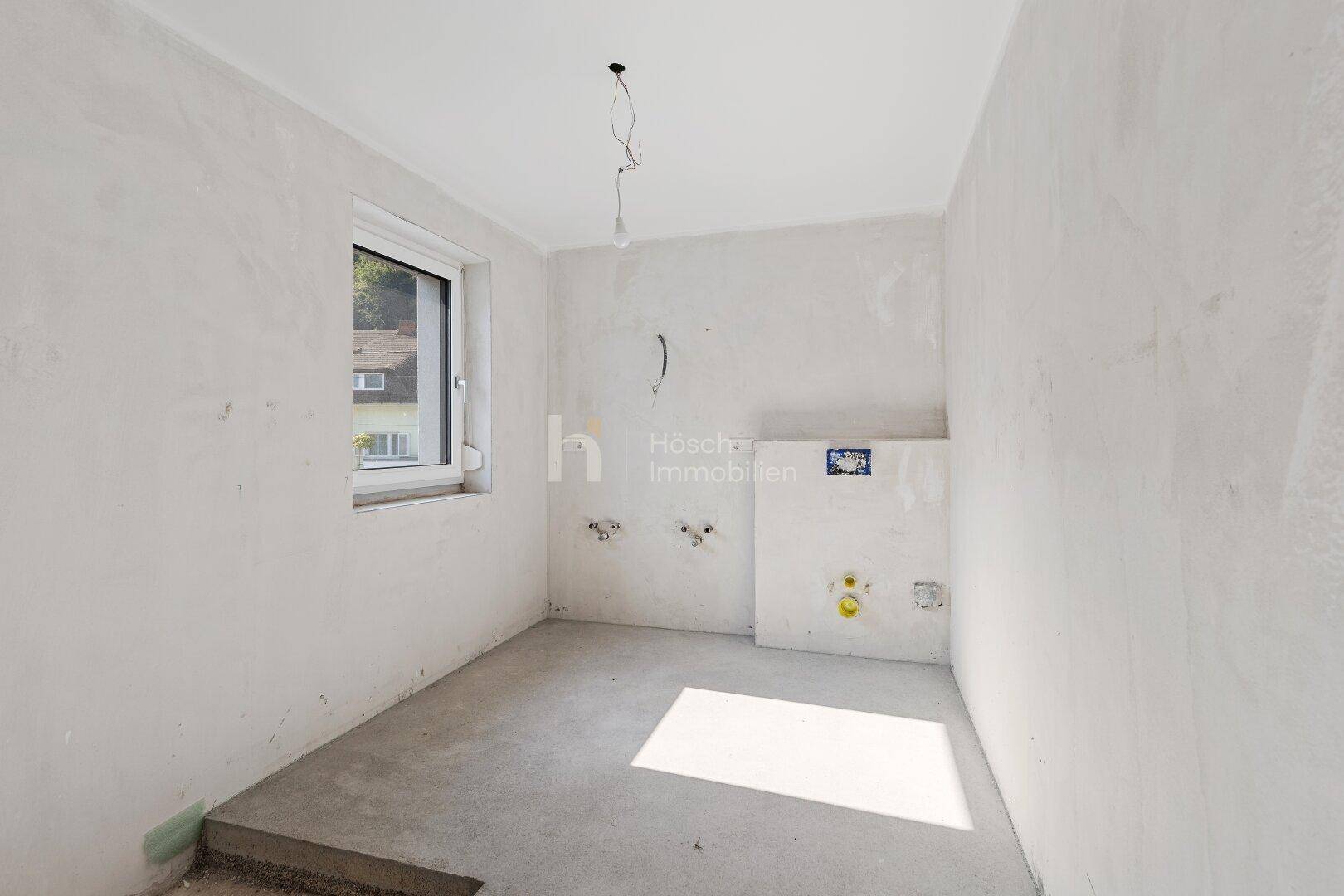 Unfertiges Badezimmer mit Fenster, sichtbaren Anschlüssen und Rohbau-Boden.