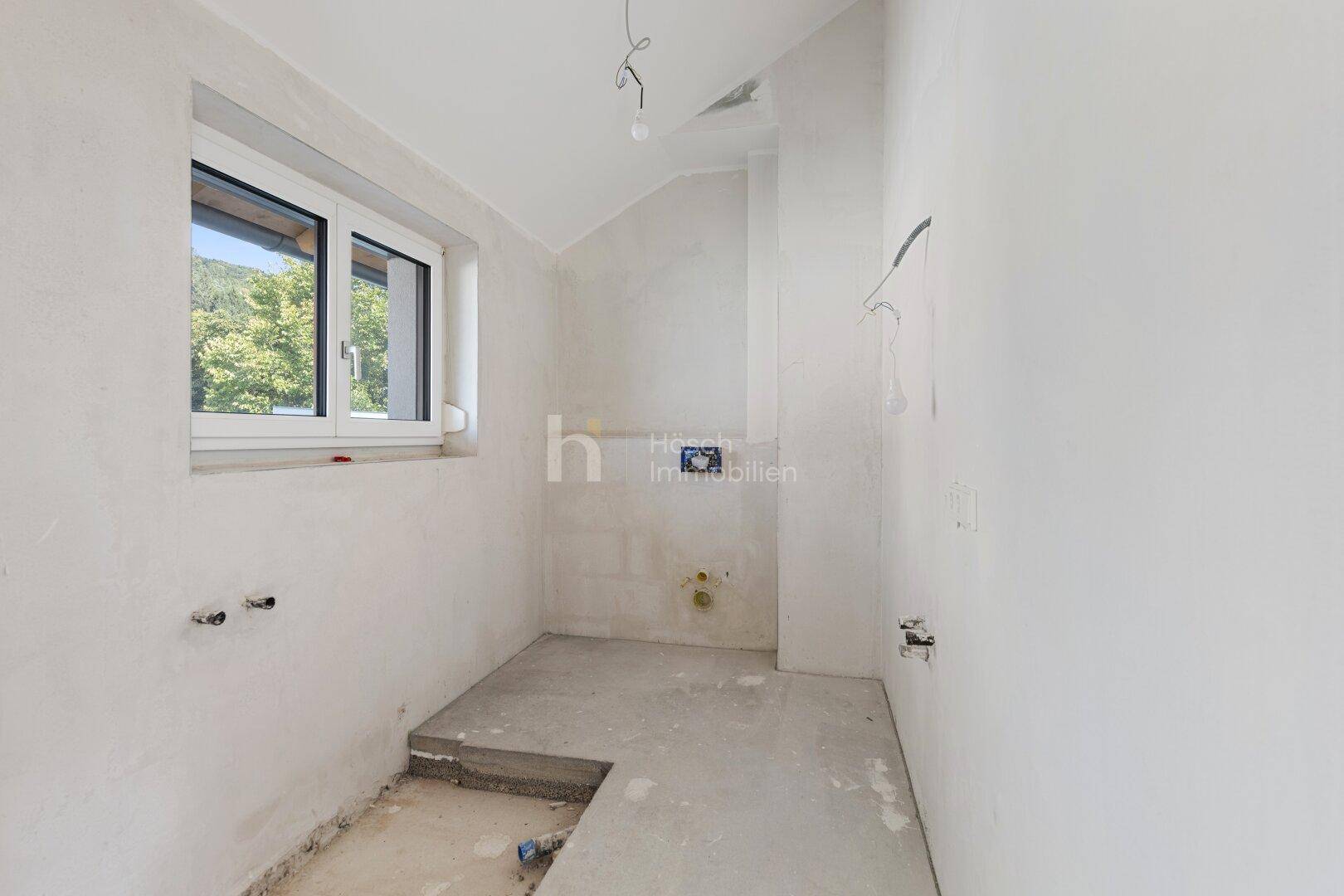 Unfertiges Badezimmer im Dachgeschoss mit Fenster, sichtbaren Anschlüssen und Rohbau-Boden.