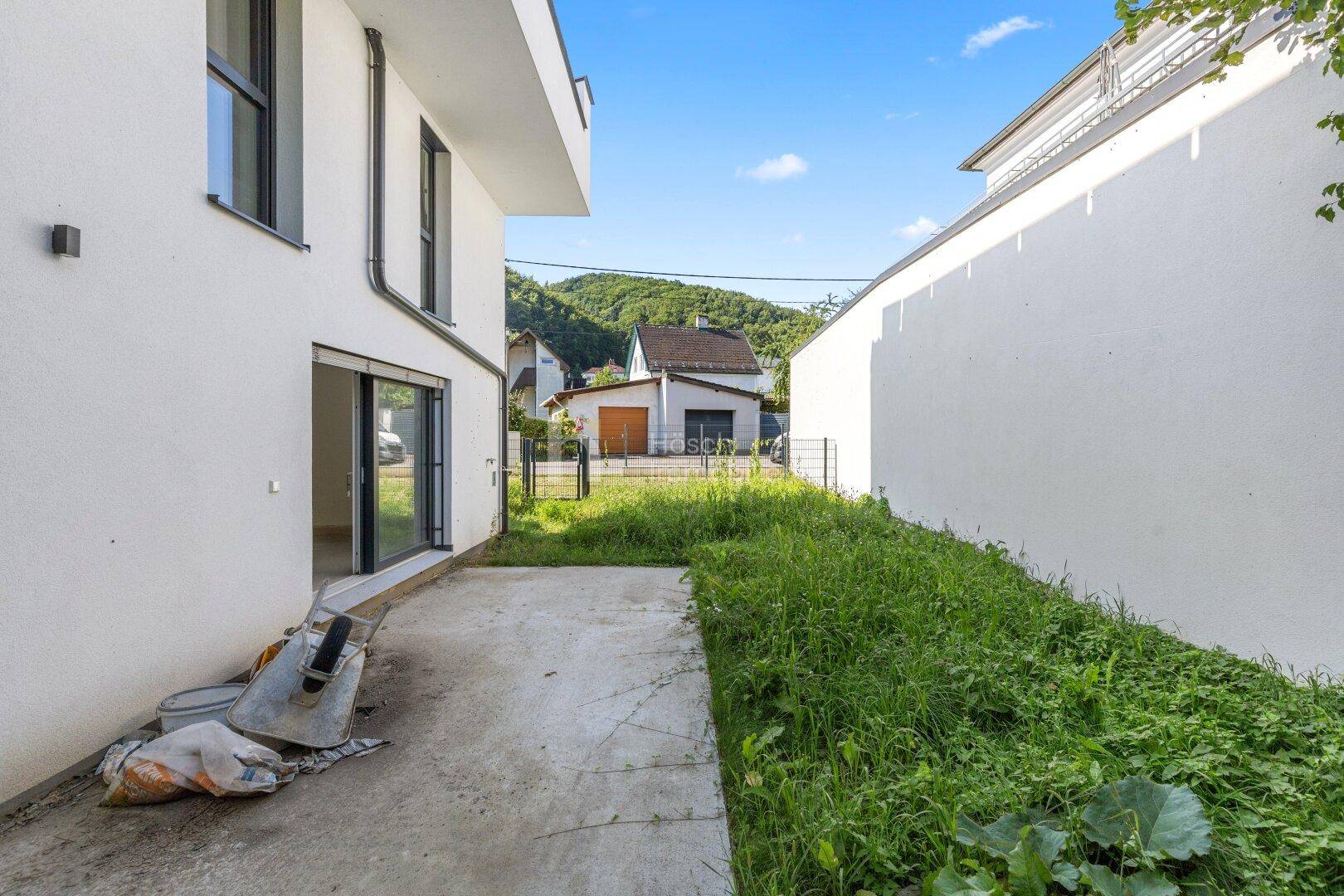 Hinterhofbereich eines Neubaus mit unfertiger Terrasse und wild wachsender Grünfläche.