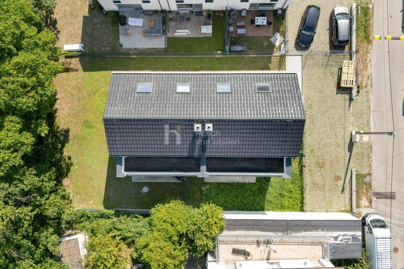 Draufsicht auf ein modernes Haus mit dunklem Satteldach und Dachfenstern, umgeben von Grünflächen.