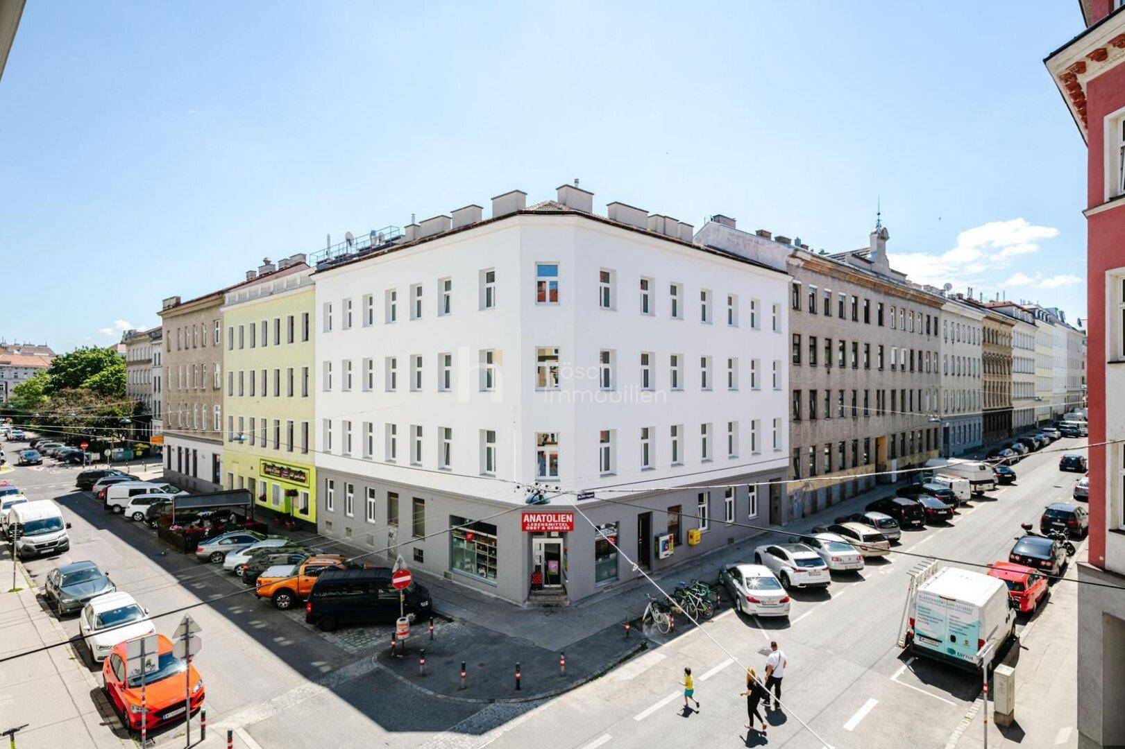 Eckgebäude in einer belebten Stadtstraße mit vielen Fenstern und Geschäften im Erdgeschoss.