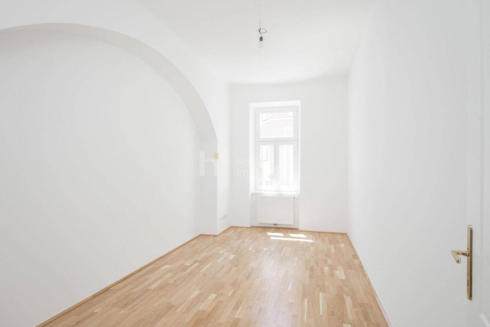 Helles Zimmer mit Parkettboden, einem Fenster und einem dekorativen Bogen in der Wand.