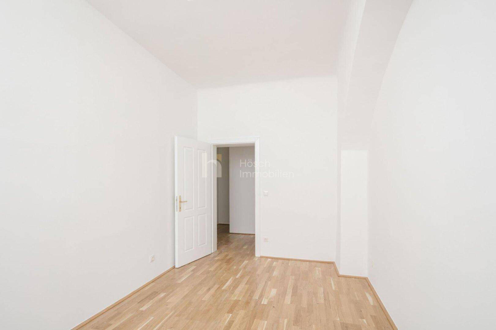 Leeres Zimmer mit Parkettboden und einer geöffneten Tür, die den Blick in einen weiteren Raum freigibt.