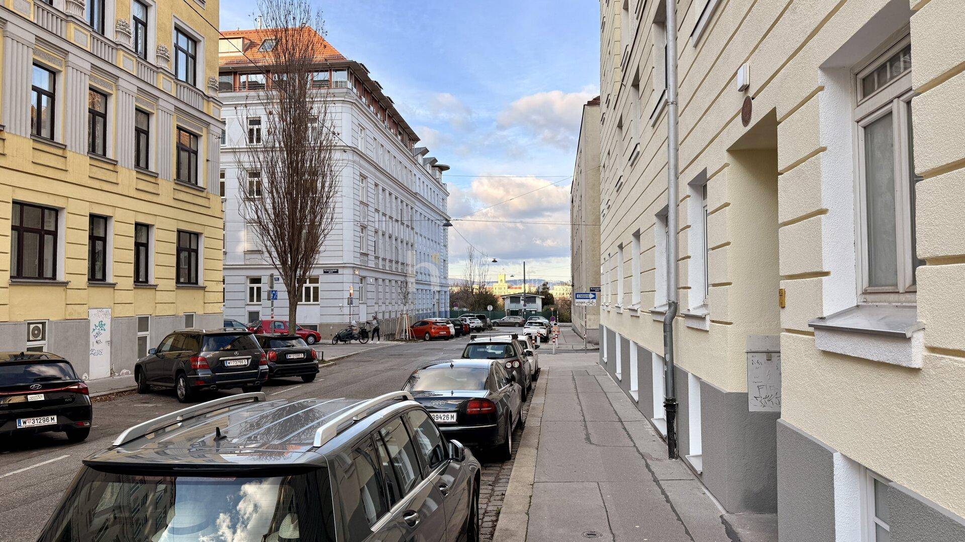 Eine belebte Stadtstraße mit parkenden Autos und mehrstöckigen Wohngebäuden unter blauem Himmel.