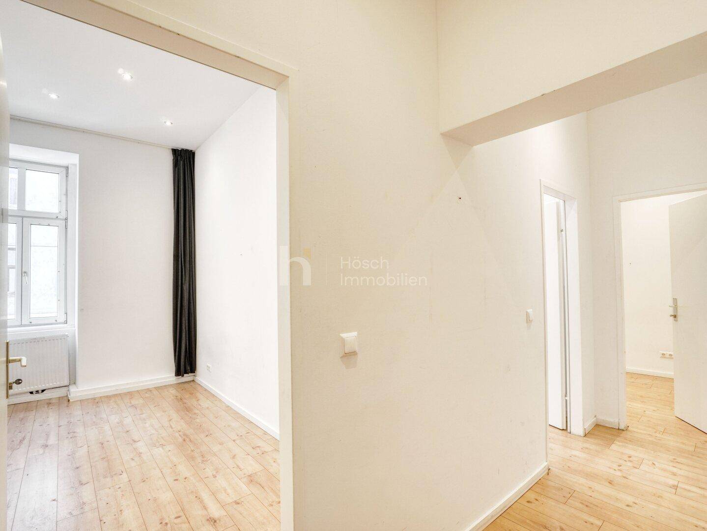 Heller Flur mit Holzboden, der zu einem Zimmer mit großem Fenster und dunklen Vorhängen führt.