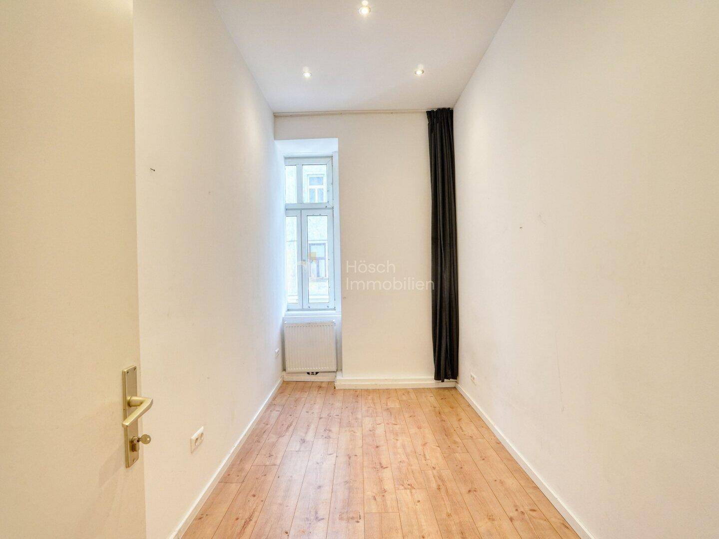 Leeres Zimmer mit Holzboden, einem hohen Fenster und dunklen Vorhängen, bereit zur individuellen Gestaltung.