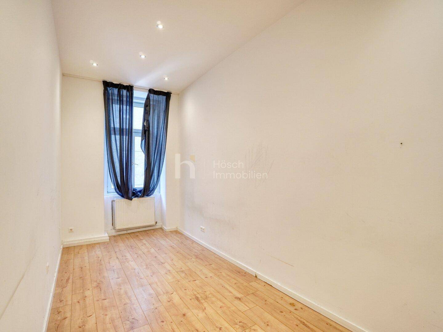 Unmöbliertes Zimmer mit Holzboden, einem Fenster mit dunklen Vorhängen und Einbaustrahlern an der Decke.