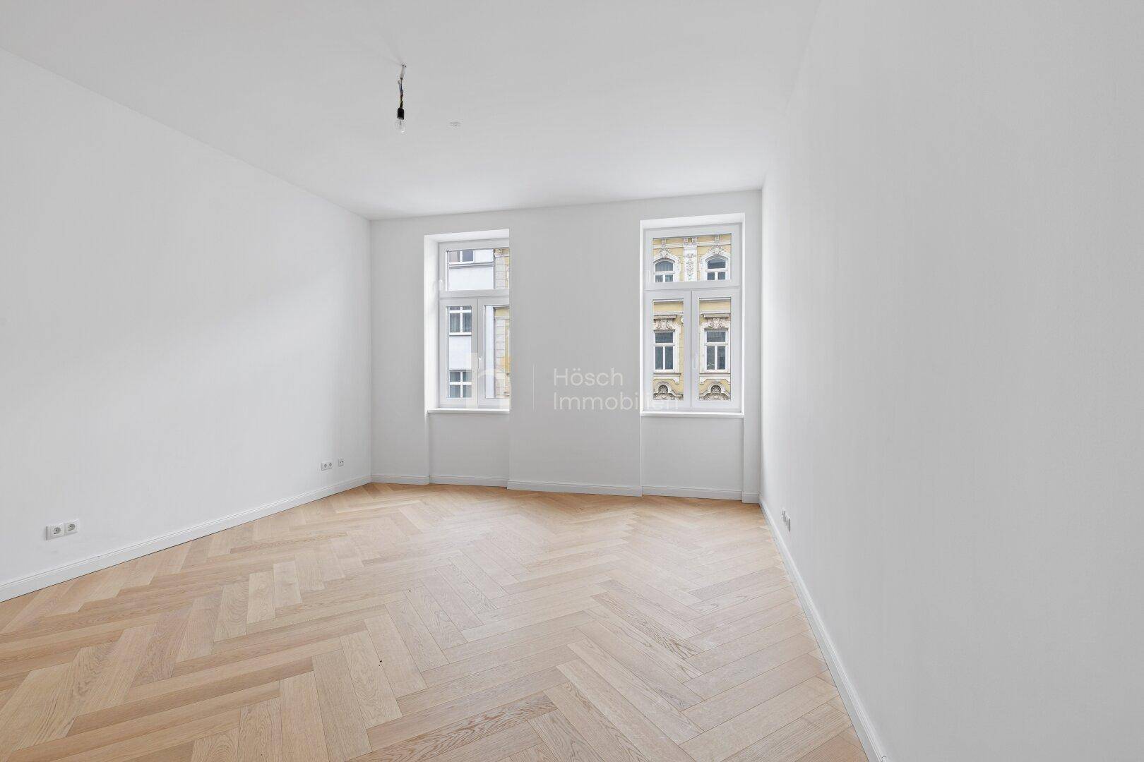 Helles, leeres Zimmer mit Fischgrätparkett und zwei Fenstern, die einen Blick nach draußen bieten.