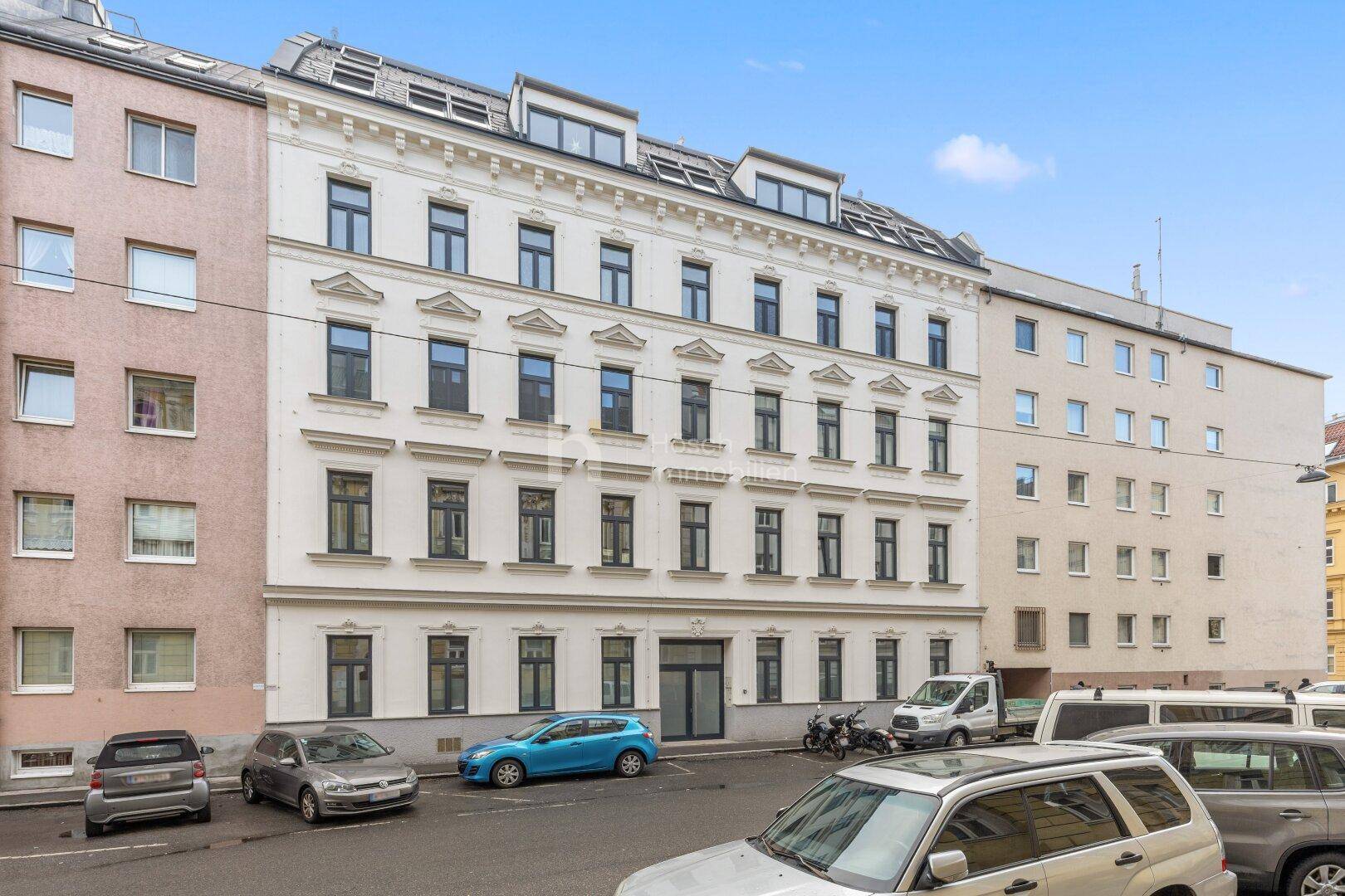 Klassische Altbaufassade mit vielen Fenstern und geparkten Autos davor in urbaner Umgebung.