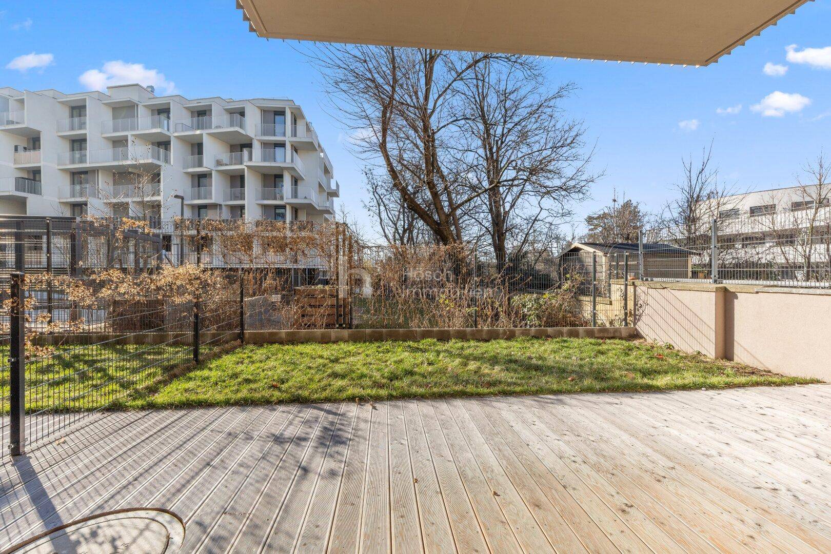 Gartenbereich mit Rasenfläche und Holzterrasse, umgeben von einem Zaun und Gebäuden.