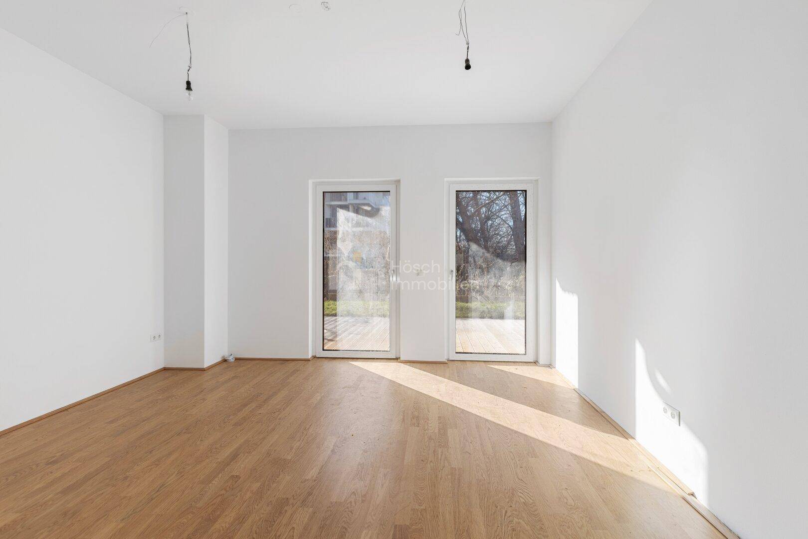 Lichtdurchfluteter Wohnraum mit Parkettboden und zwei Türen, die zu einem Außenbereich führen.