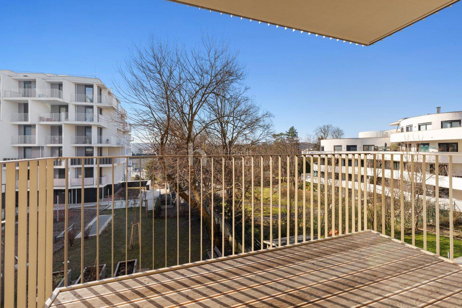 Balkon mit Holzboden und Blick auf eine grüne Gartenanlage sowie moderne Wohngebäude.