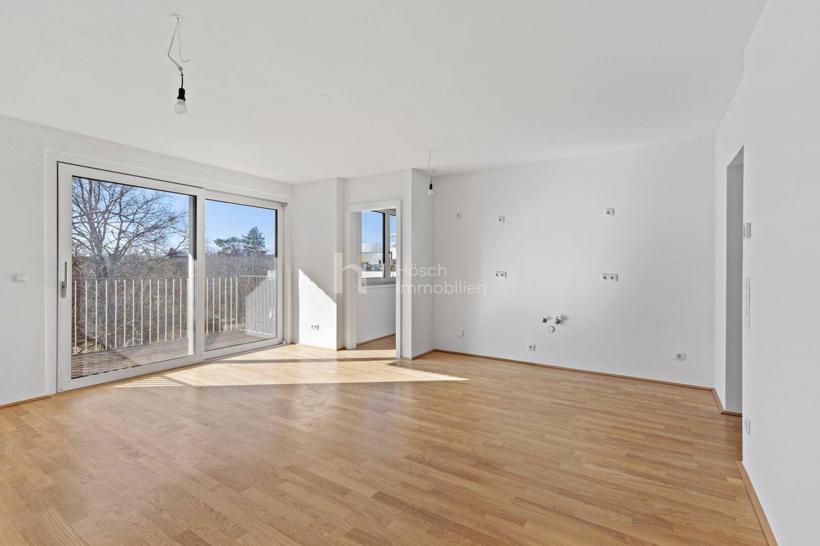 Heller, leerer Wohnraum mit Parkettboden, großen Fenstern und Zugang zu einem Balkon.