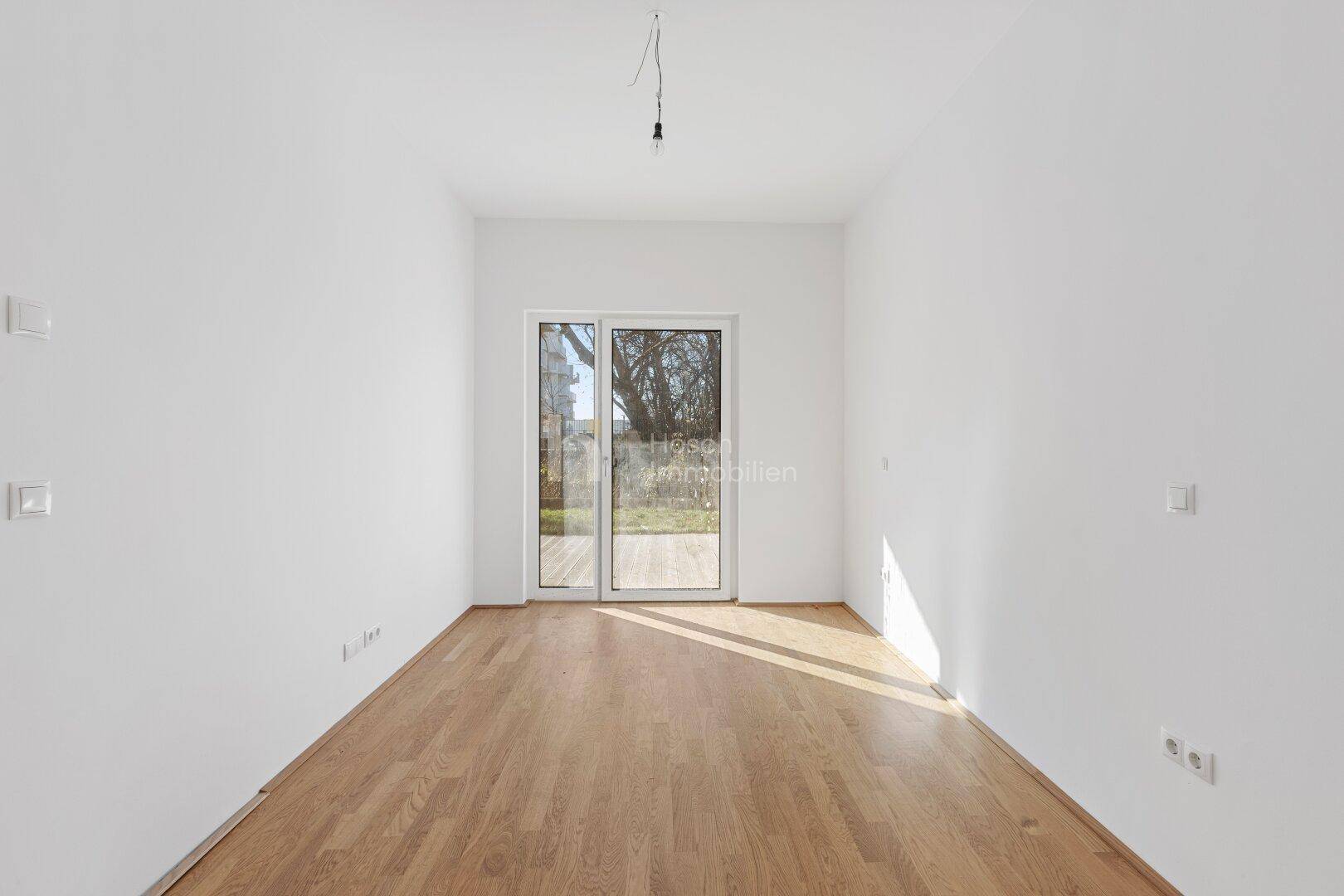 Geräumiger, heller Raum mit Holzboden und einer großen Glastür, die den Blick ins Grüne freigibt.