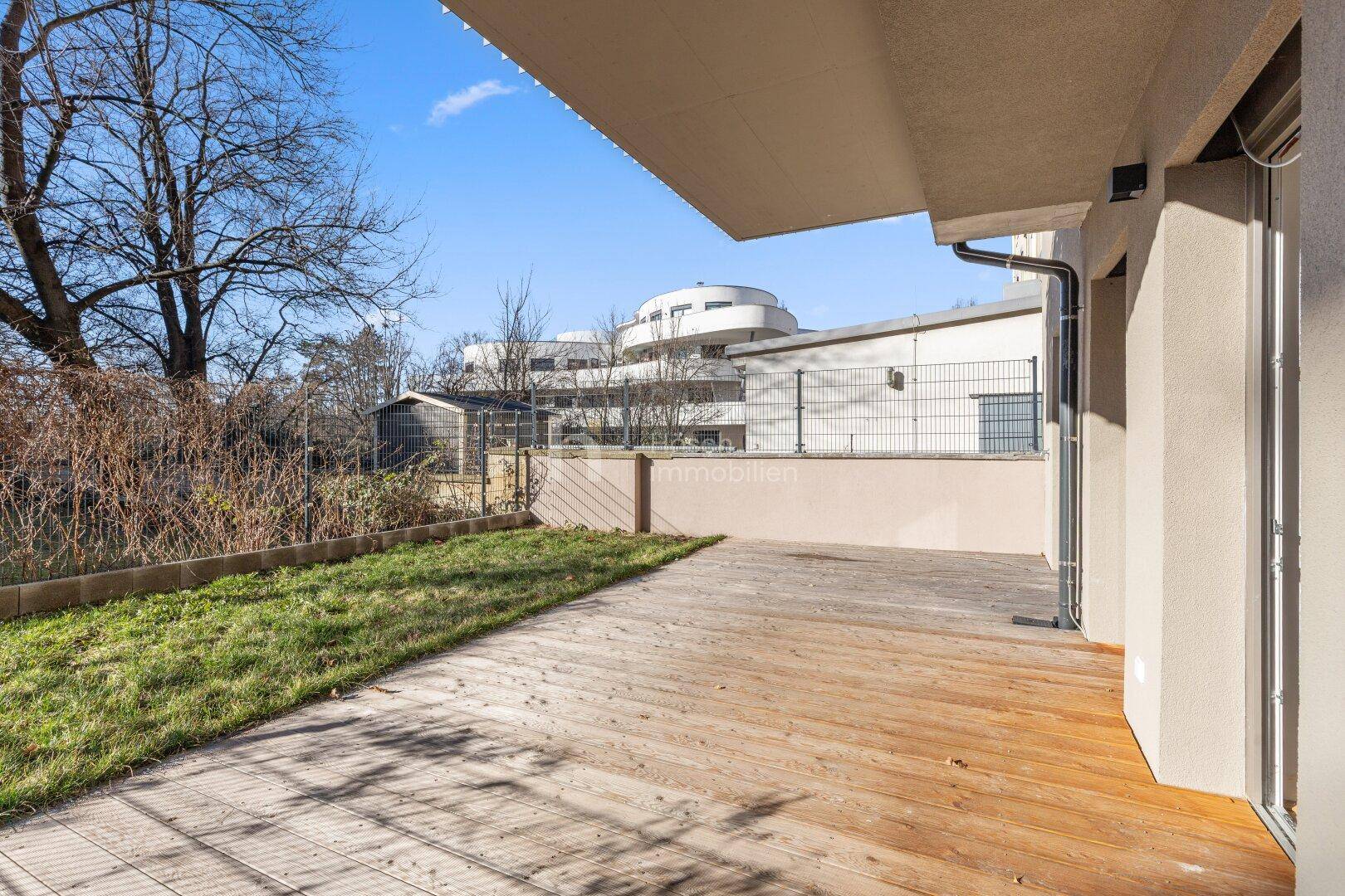 Sonniger Außenbereich mit Holzterrasse und kleinem Garten, umgeben von moderner Architektur.