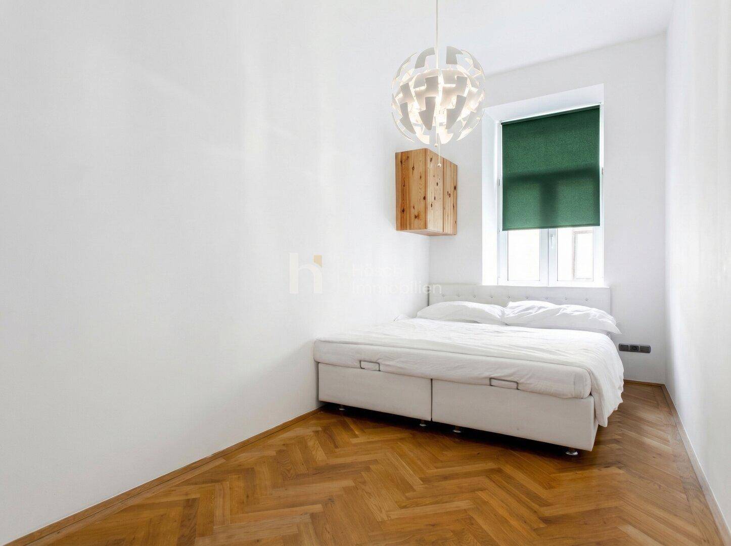Helles Schlafzimmer mit weißem Bett, Holzparkett und einem Fenster mit grünem Rollo.