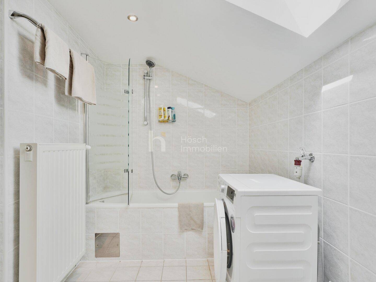 Gefliestes Badezimmer mit Badewanne, Duschvorrichtung und Waschmaschine unter der Dachschräge.