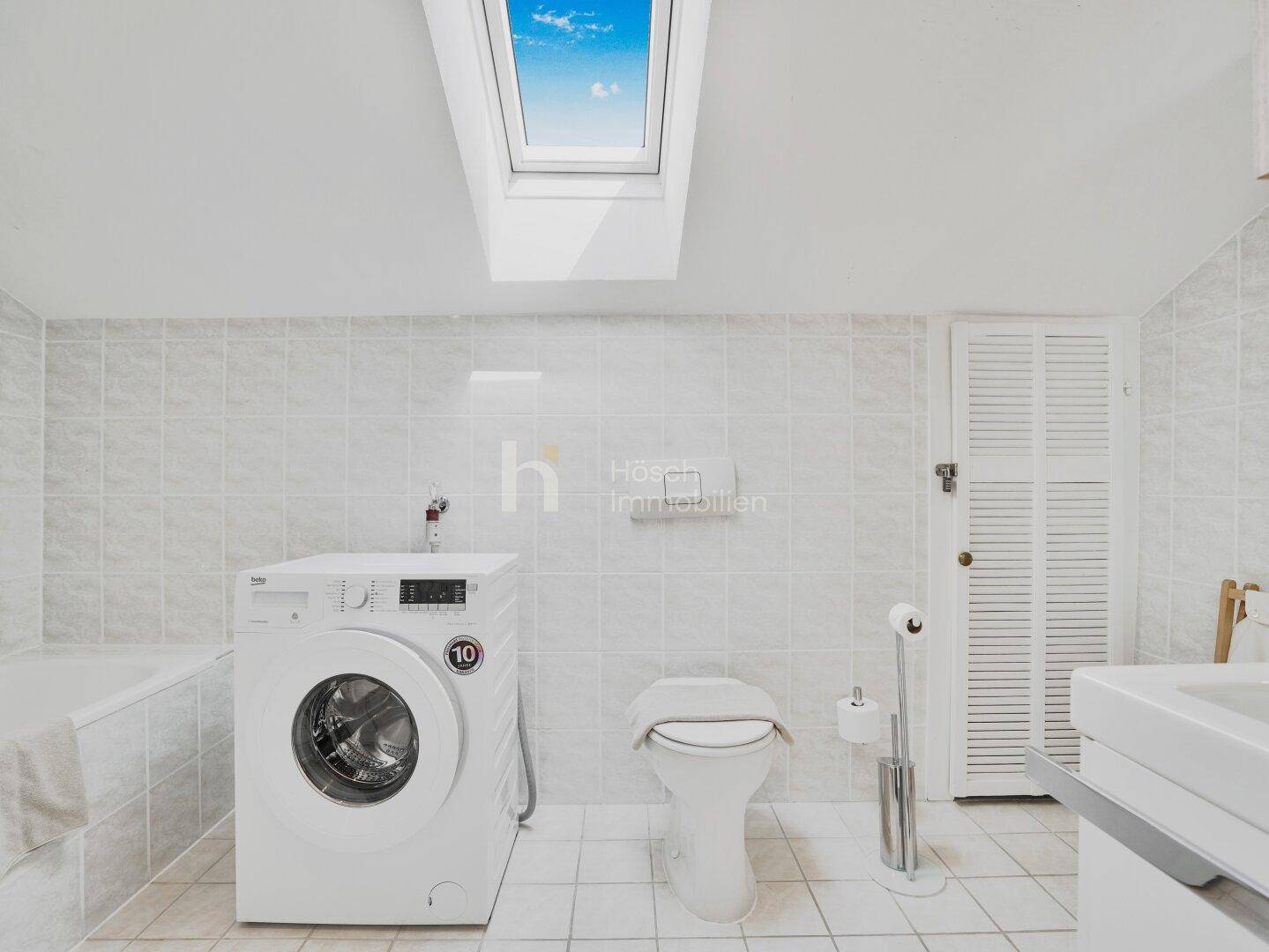 Badezimmer mit Dachfenster, WC, Waschmaschine und hellen Fliesen.