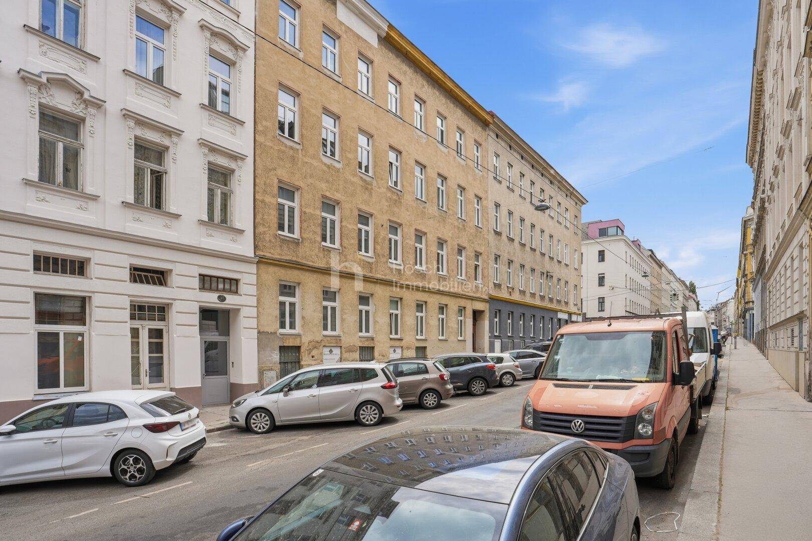 Historische Straßenansicht eines Altbaus mit parkenden Autos und umliegenden Gebäuden.