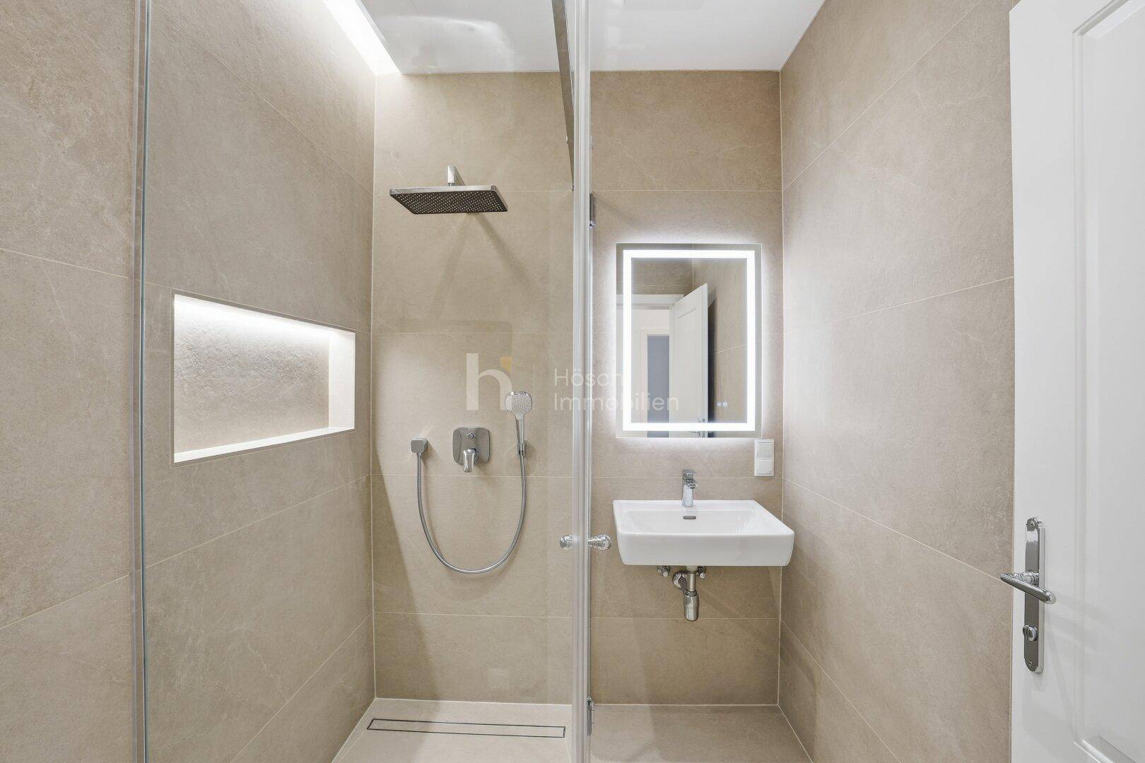 Modernes Badezimmer mit beigefarbenen Fliesen, begehbarer Dusche und beleuchtetem Spiegel.