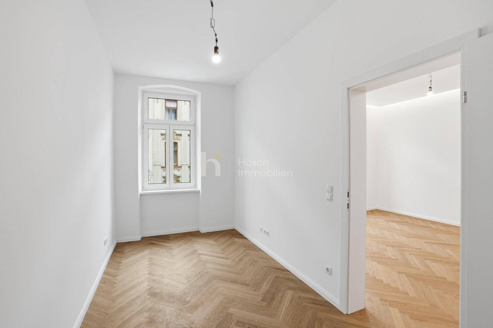 Gemütliches Zimmer mit Fischgrätparkett, einem Fenster und Zugang zu einem angrenzenden Raum.