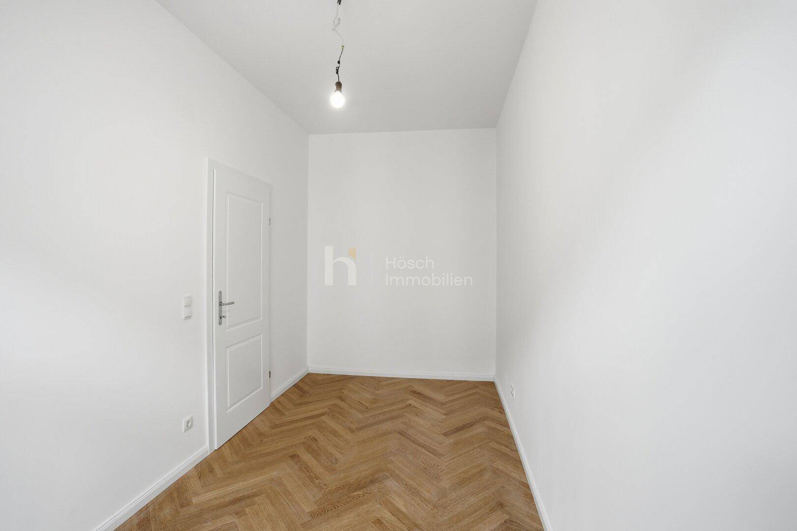 Helles Zimmer mit Fischgrätparkett und einer weißen Tür, ideal als Schlaf- oder Arbeitszimmer.