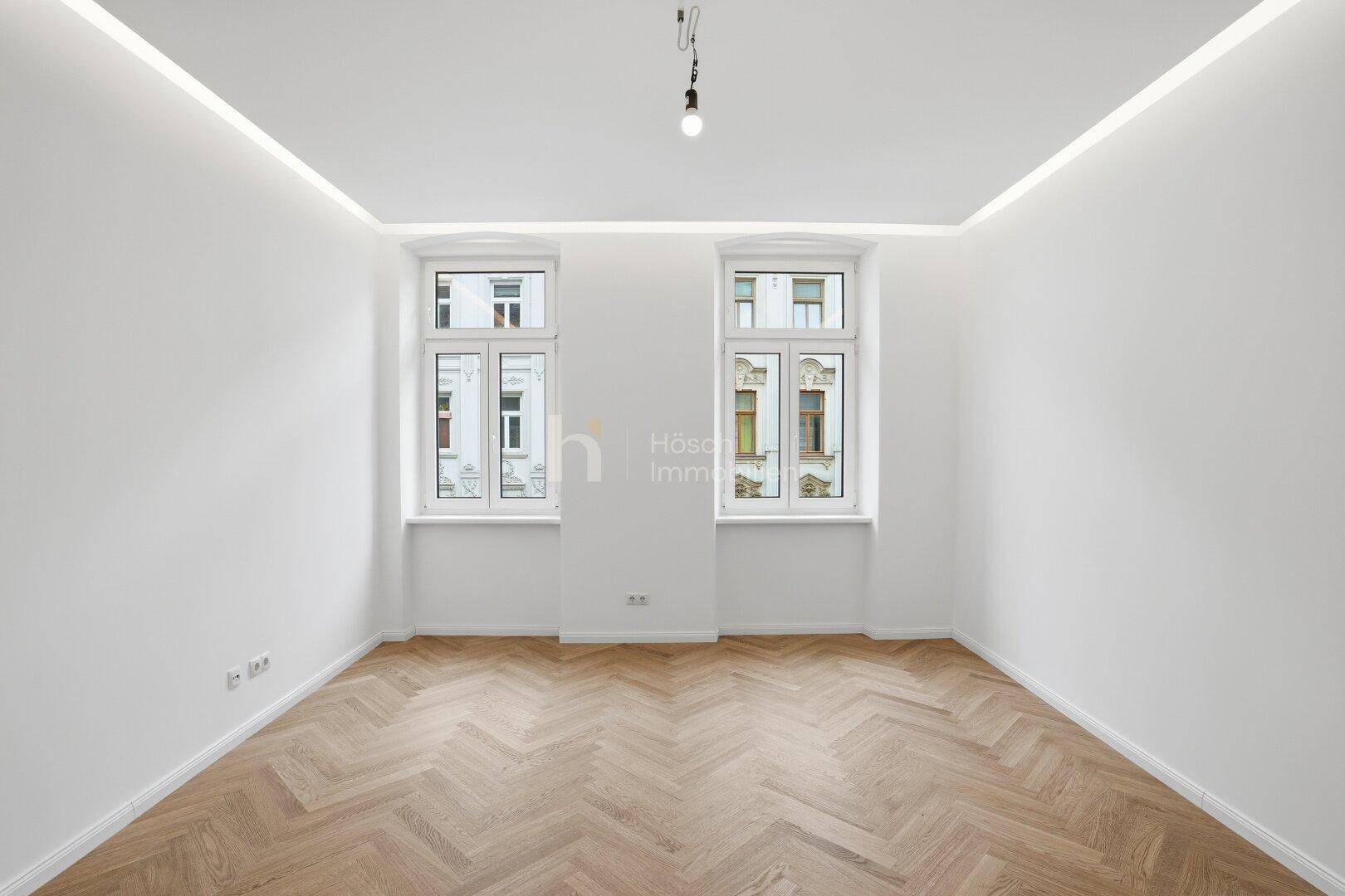 Helles Zimmer mit Fischgrätparkettboden, weißen Wänden und modernen Deckenleuchten.